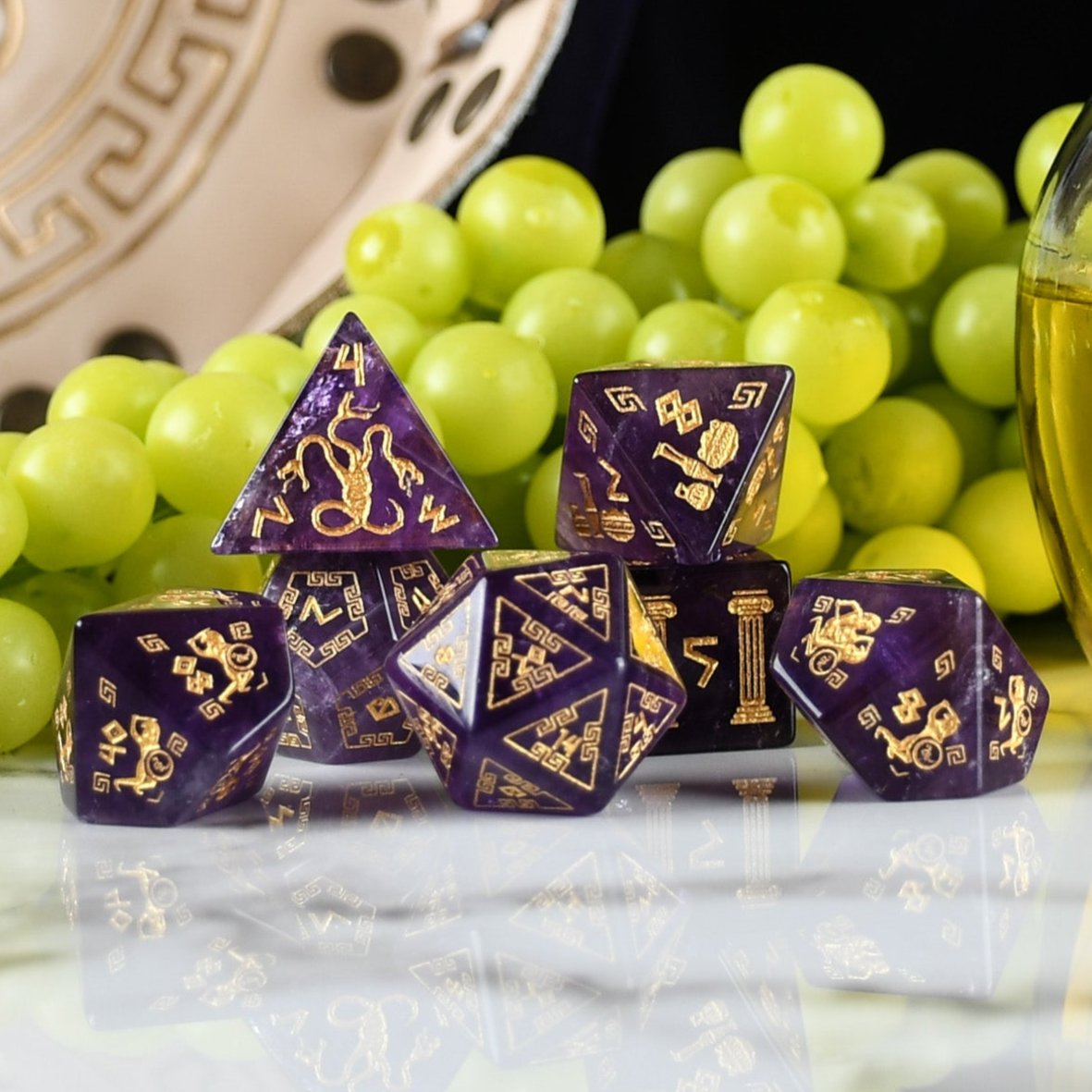 A set of purple amethyst stone dice with gold engraved designs, displayed alongside green grapes and a dnd dice tray in the background.