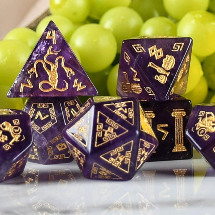 Purple polyhedral dice set with gold symbols on a reflective surface with green grapes in the background