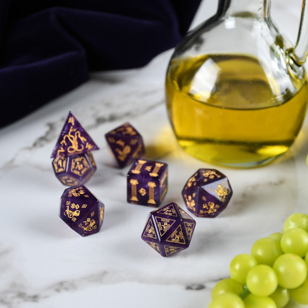 A set of purple amethyst stone dice with gold engraved designs, displayed alongside green grapes and a dnd dice tray in the background.