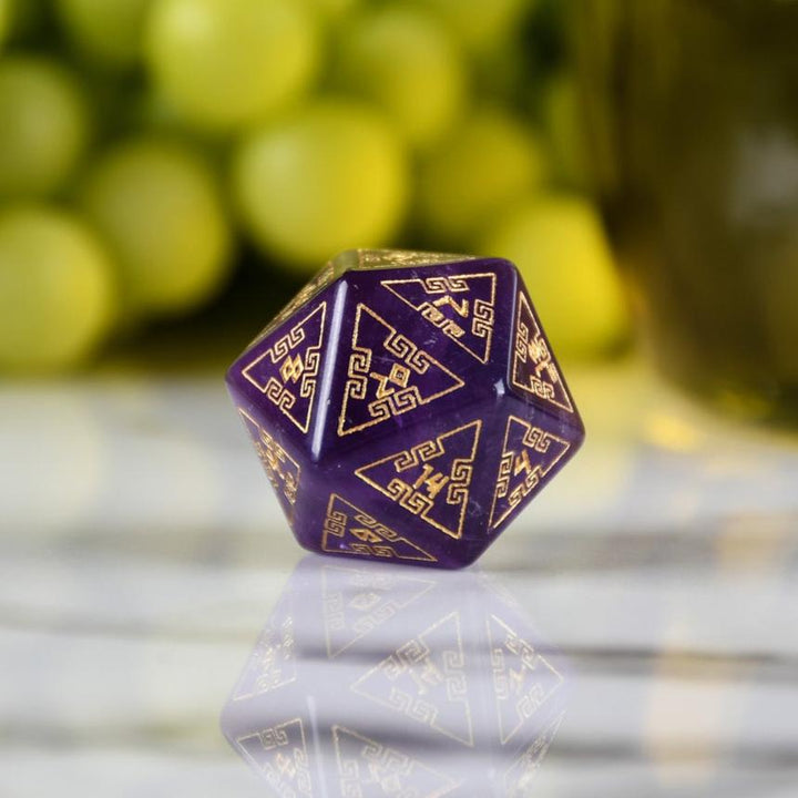A set of purple amethyst stone dice with gold engraved designs, displayed alongside green grapes and a dnd dice tray in the background.