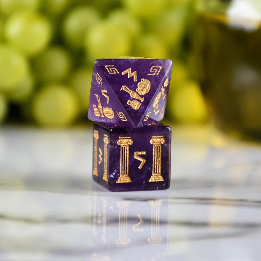 A set of purple amethyst stone dice with gold engraved designs, displayed alongside green grapes and a dnd dice tray in the background.
