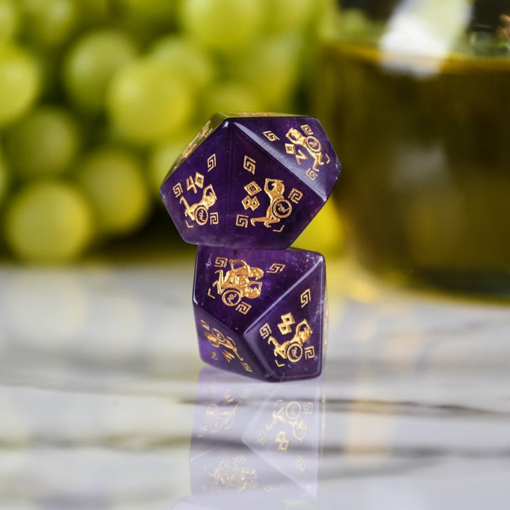 A set of purple amethyst stone dice with gold engraved designs, displayed alongside green grapes and a dnd dice tray in the background.