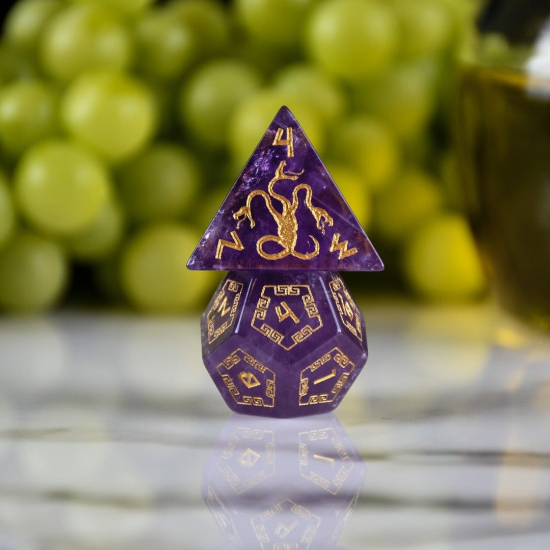 A set of purple amethyst stone dice with gold engraved designs, displayed alongside green grapes and a dnd dice tray in the background.