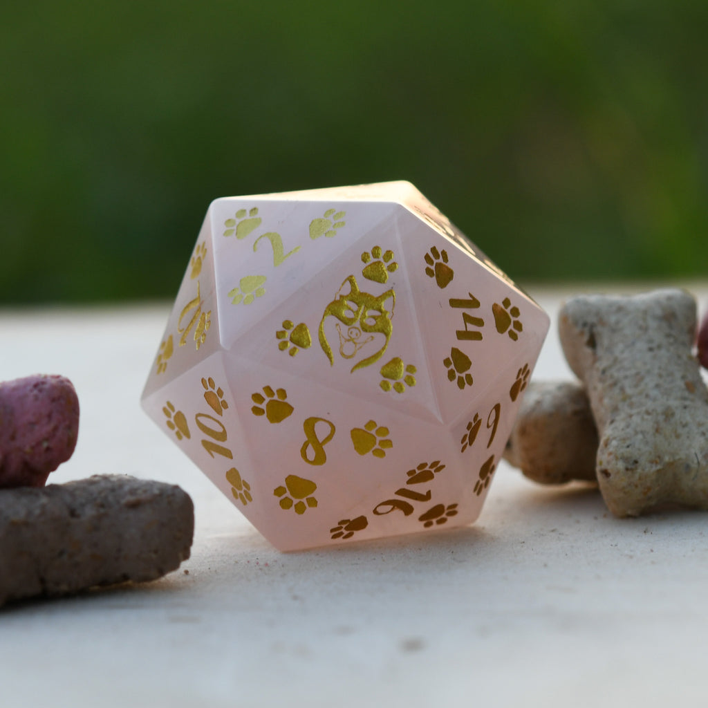 A pink rose quartz D20 dice with gold puppy paw prints and numbers engraved and painted on it, displayed next to what appears to be a dog bone.