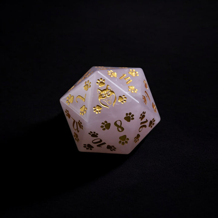 A pink rose quartz D20 dice with gold puppy paw prints and numbers engraved and painted on it