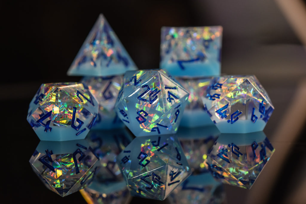 A set of blue iridescent sharp-edged, multicolored resin dice with engraved numbers, reflecting on a mirrored surface.