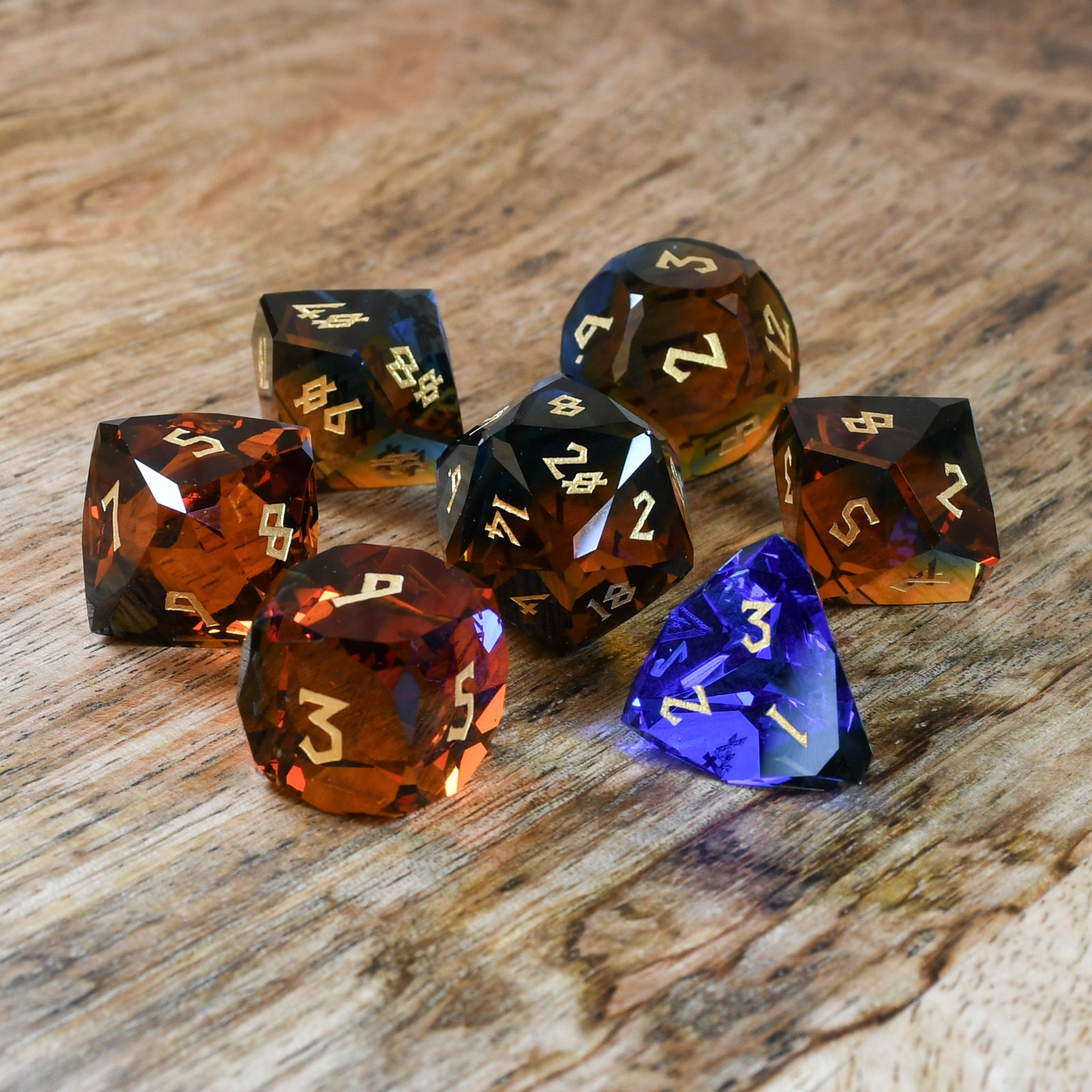 A set of multifaceted glass dice with engraved numbers, including various shapes such as a four-sided die (D4), six-sided die (D6), eight-sided die (D8), ten-sided die (D10), twelve-sided die (D12), and twenty-sided die (D20). The dice are placed on a wooden surface.