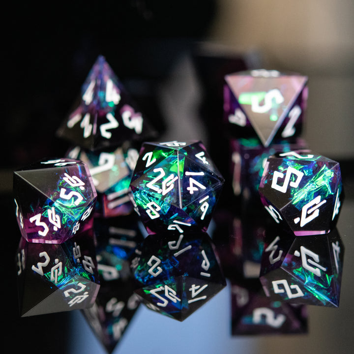 A collection of sharp-edged resin dice in black with hand-painted white numbers, featuring a metallic foil inside with hints of purple, green, and blue.
