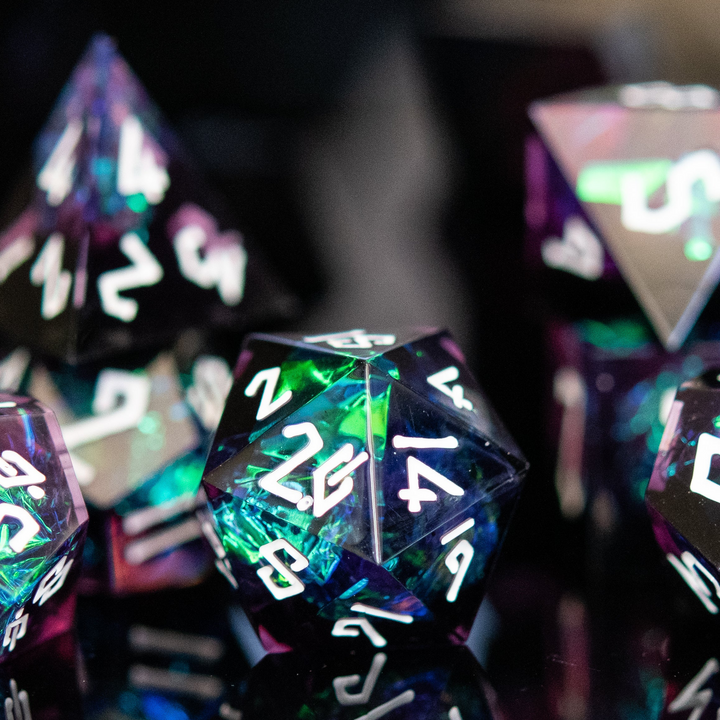 A collection of sharp-edged resin dice in black with hand-painted white numbers, featuring a metallic foil inside with hints of purple, green, and blue.
