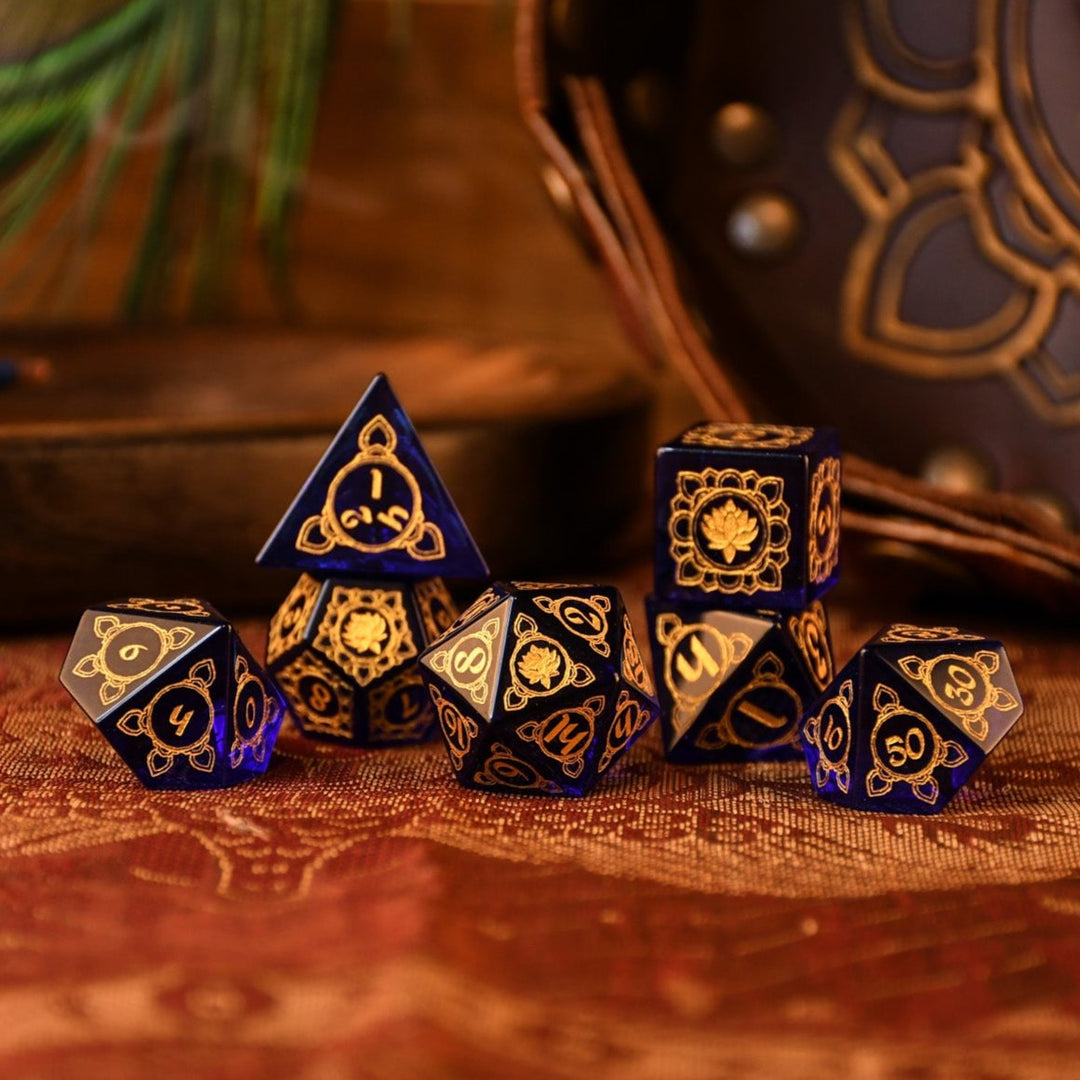 A set of hand-carved blue glass dice with golden designs, including a D4, D6, D8, D10, D12, and D20, displayed on a textured surface.