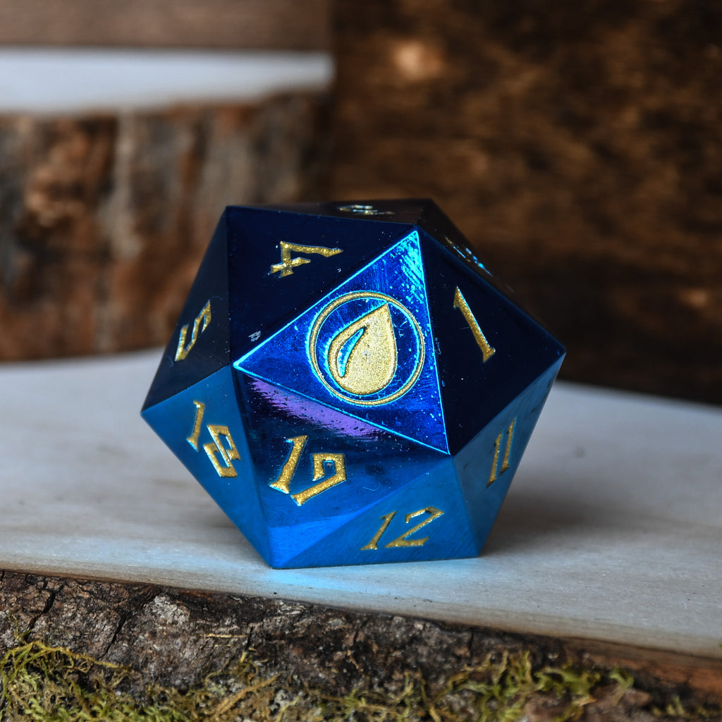 A solid-metal spindown life counter d20 dice, predominantly blue with gold numbers and a gold-colored logo on one face, placed on a white surface with a wood grain background.