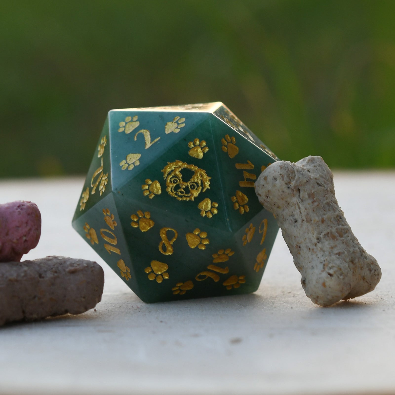 A green aventurine D20 dice with gold puppy paw prints and engravings, displayed next to a bone-shaped object.