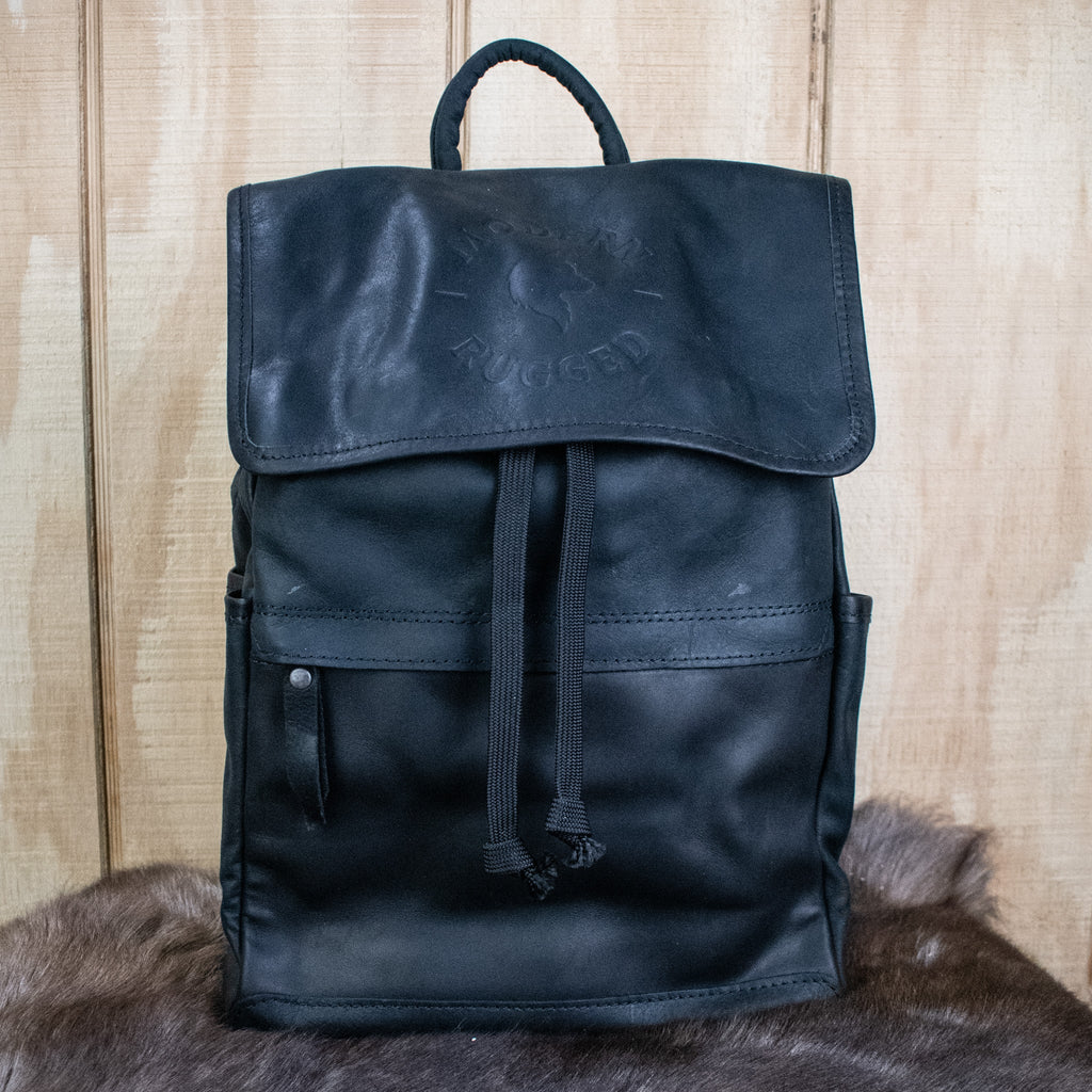 A black leather hide backpack with brass zippers and a drawstring top, displayed against a wooden background.