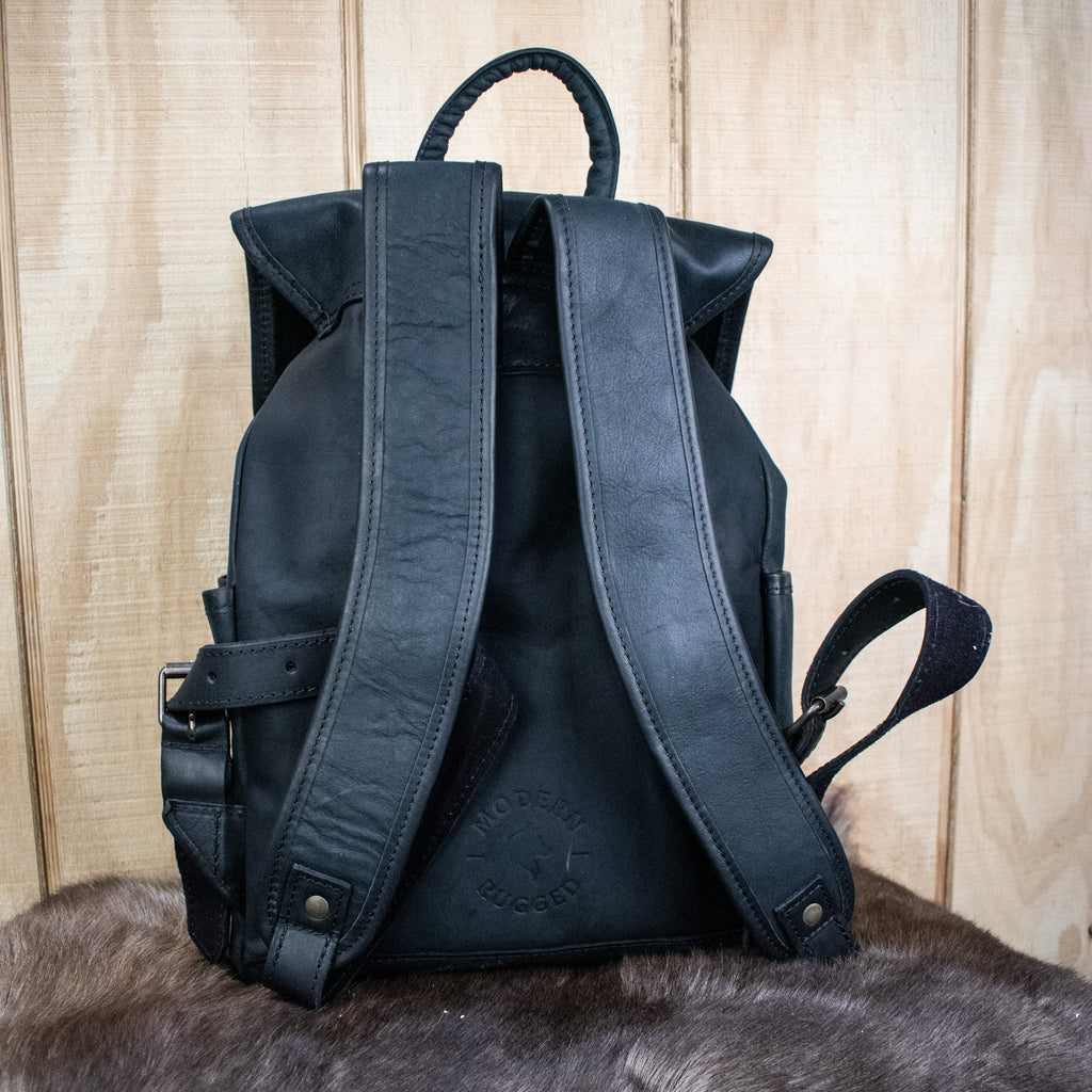A black leather hide backpack with brass zippers and a drawstring top, displayed against a wooden background.