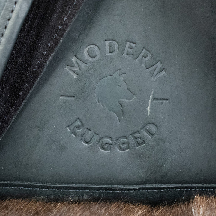 A black leather hide backpack with brass zippers and a drawstring top, displayed against a wooden background.