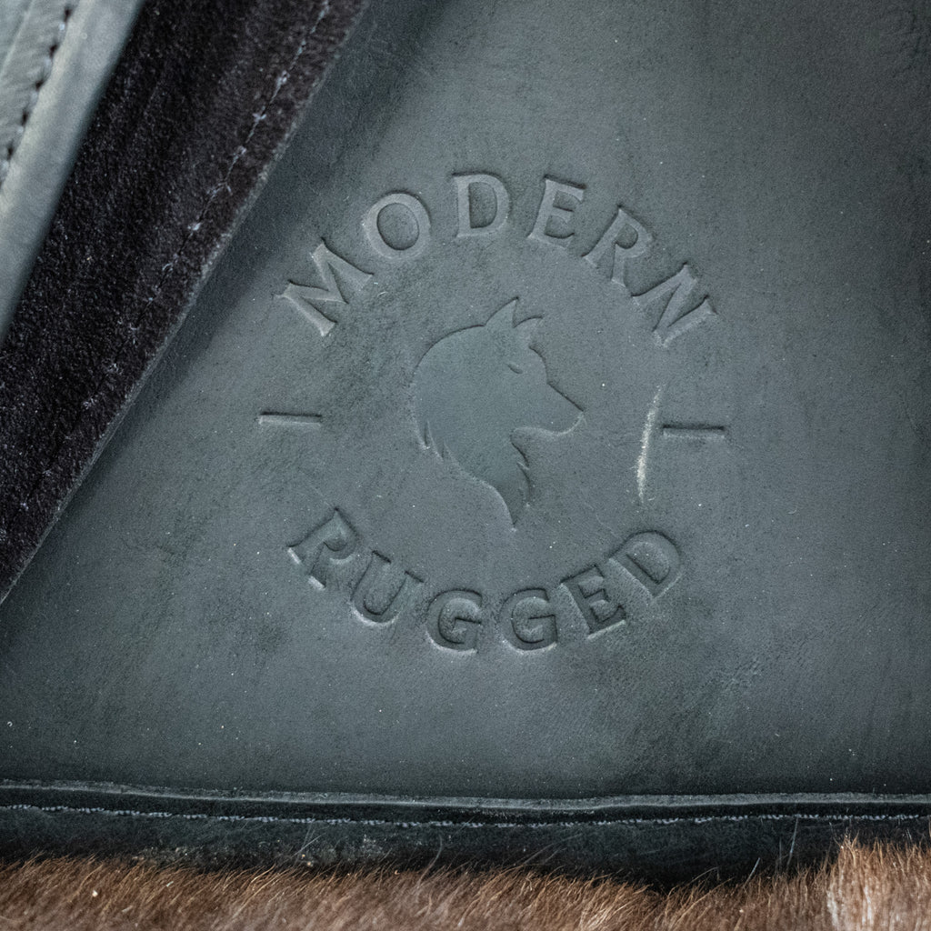 A black leather hide backpack with brass zippers and a drawstring top, displayed against a wooden background.