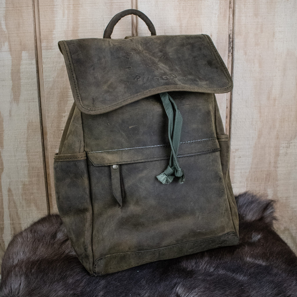 A weathered olive green leather hide backpack with a front zipper pouch, brass zippers, and padded straps, displayed on a wooden surface.