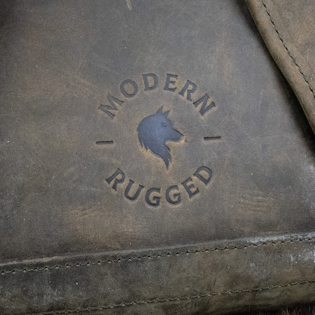 A weathered olive green leather hide backpack with a front zipper pouch, brass zippers, and padded straps, displayed on a wooden surface.