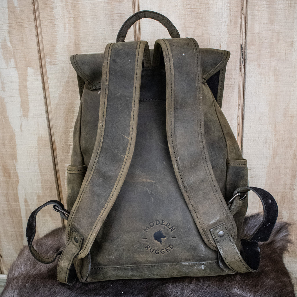 A weathered olive green leather hide backpack with a front zipper pouch, brass zippers, and padded straps, displayed on a wooden surface.
