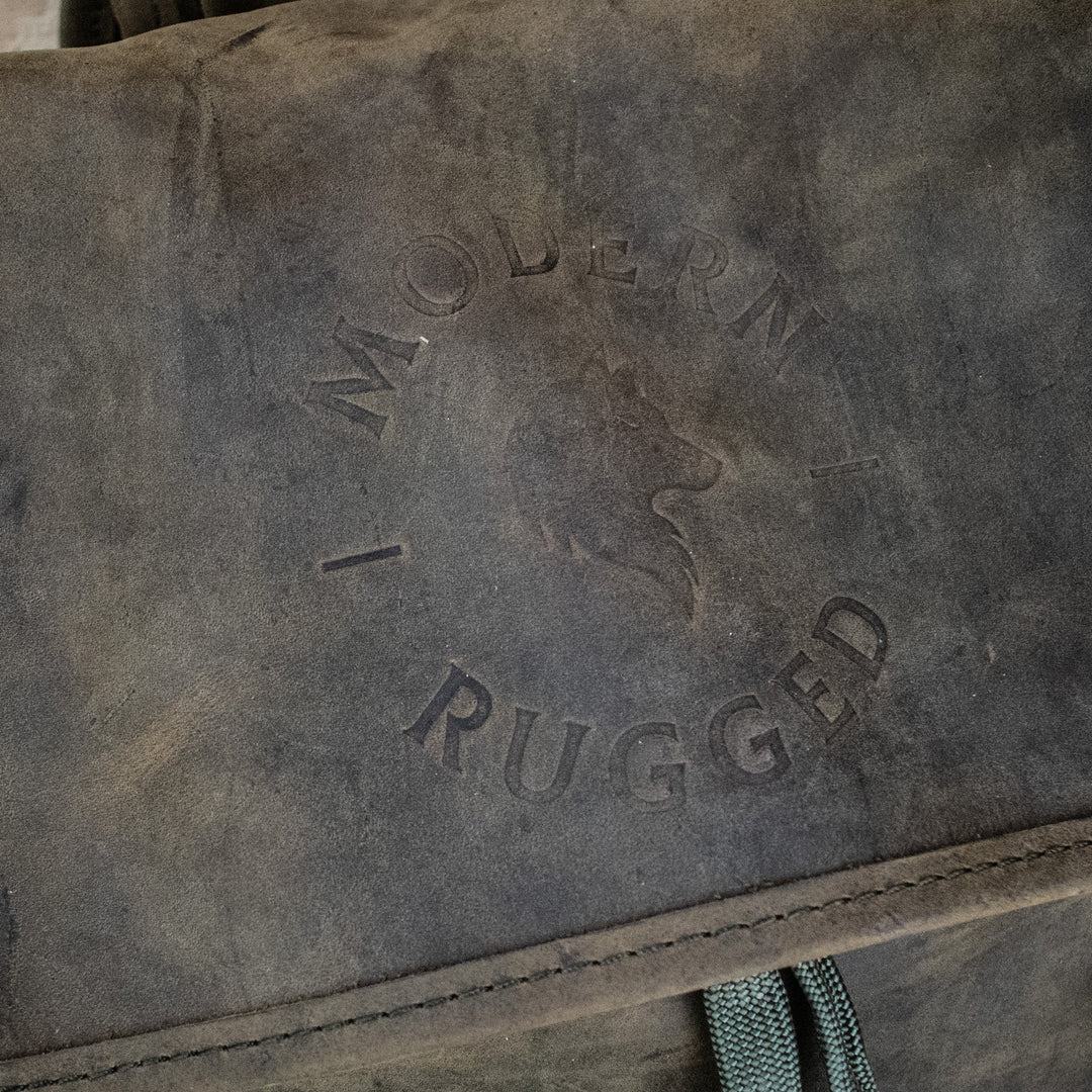 A weathered olive green leather hide backpack with a front zipper pouch, brass zippers, and padded straps, displayed on a wooden surface.