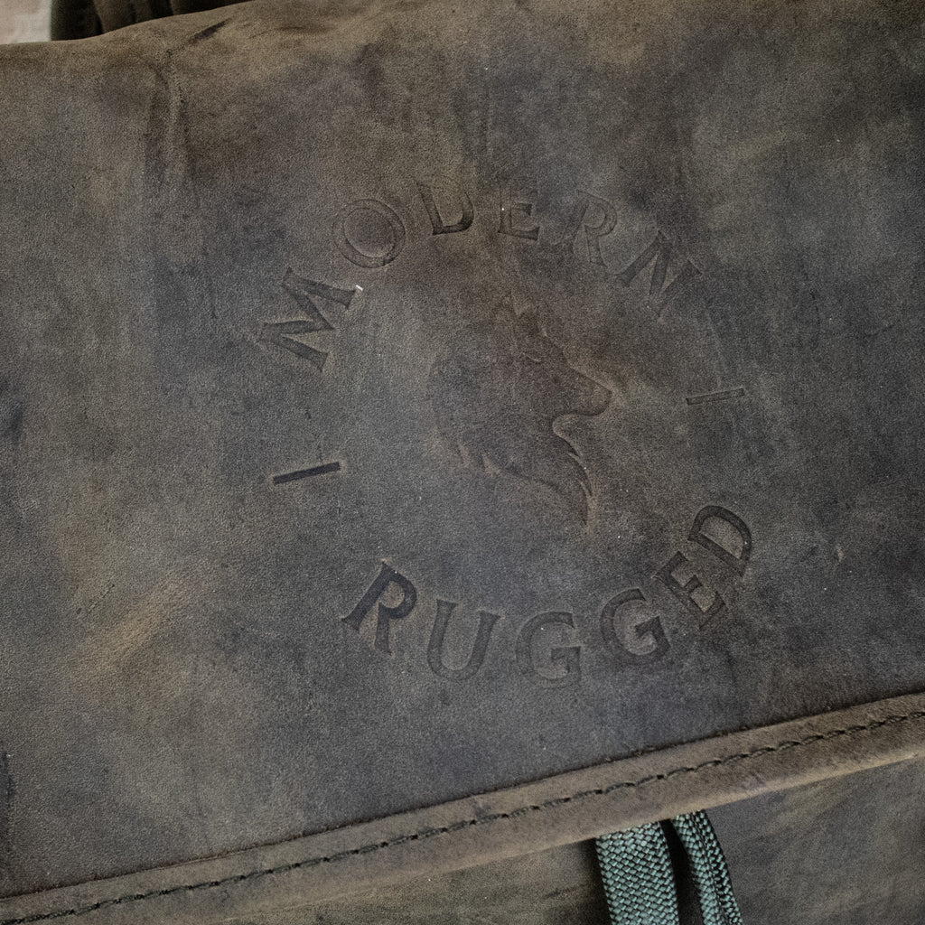 A weathered olive green leather hide backpack with a front zipper pouch, brass zippers, and padded straps, displayed on a wooden surface.