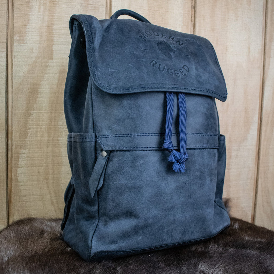 A navy-colored leather backpack with a front zipper pouch and two side pockets, featuring brass zippers and a drawstring top.