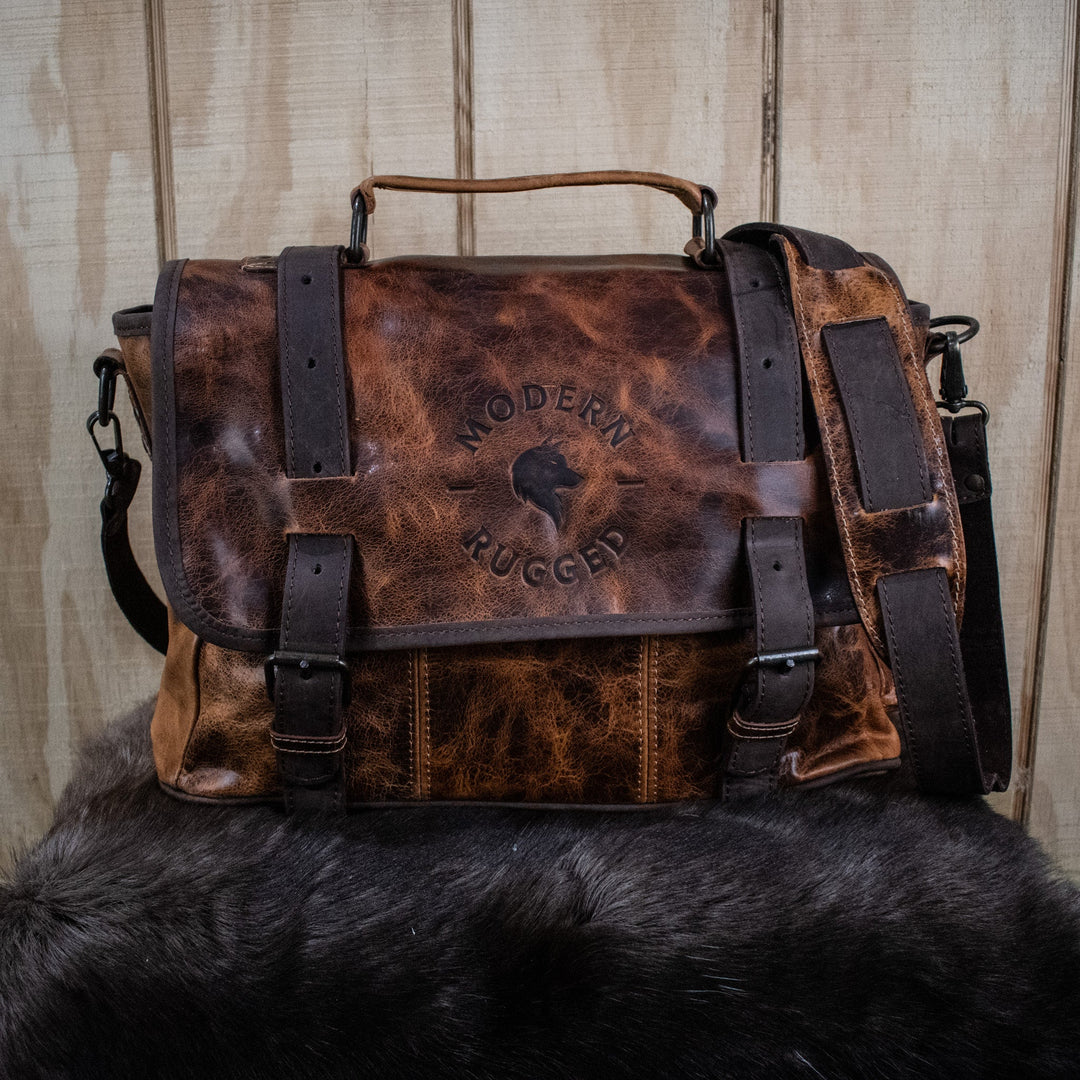A brown leather laptop bag with brass zippers and buckles, featuring the text &