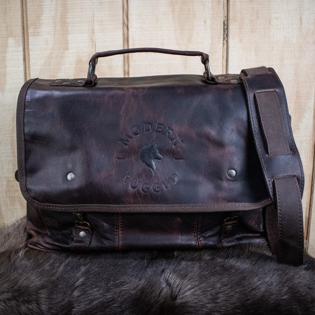 A brown leather laptop bag with brass zippers and clasps, featuring a embossed logo 'MODERN RUGGED' on the front, set against a wooden backdrop.