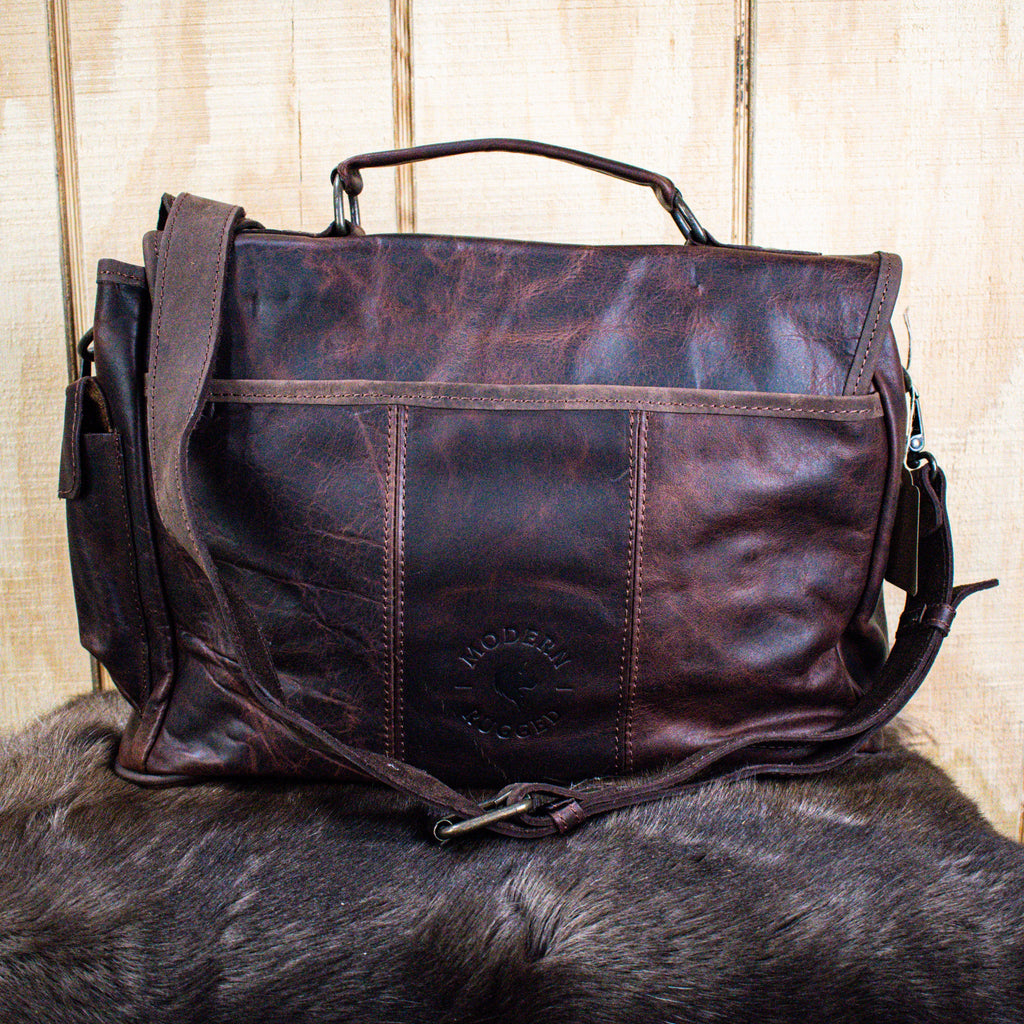 A brown leather laptop bag with brass zippers and clasps, featuring a embossed logo 'MODERN RUGGED' on the front, set against a wooden backdrop.