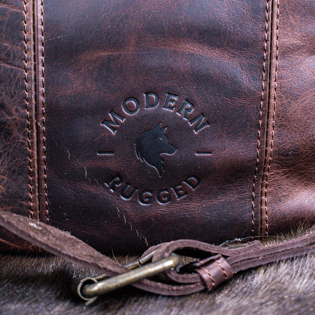 A brown leather laptop bag with brass zippers and clasps, featuring a embossed logo 'MODERN RUGGED' on the front, set against a wooden backdrop.