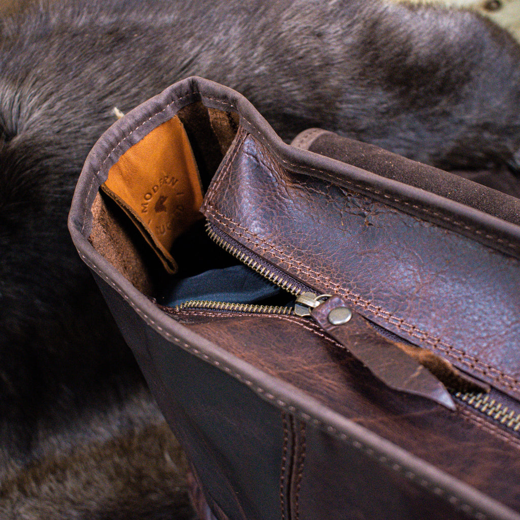 A brown leather laptop bag with brass zippers and clasps, featuring a embossed logo 'MODERN RUGGED' on the front, set against a wooden backdrop.