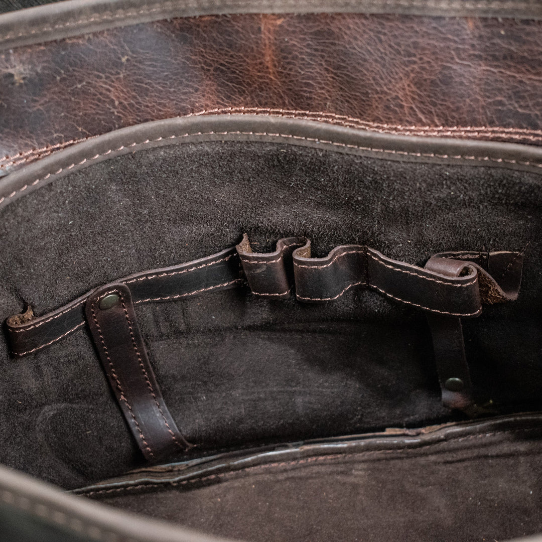 A brown leather laptop bag with brass zippers and clasps, featuring a embossed logo &