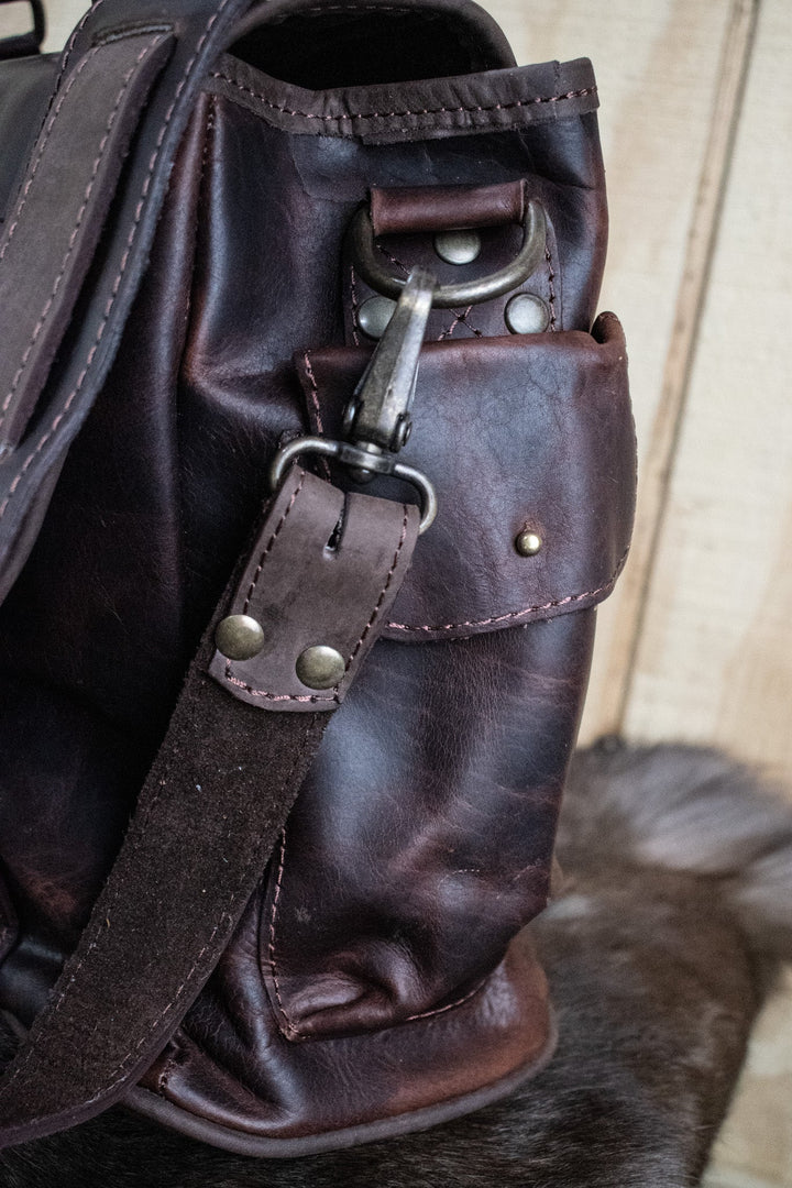 A brown leather laptop bag with brass zippers and clasps, featuring a embossed logo &