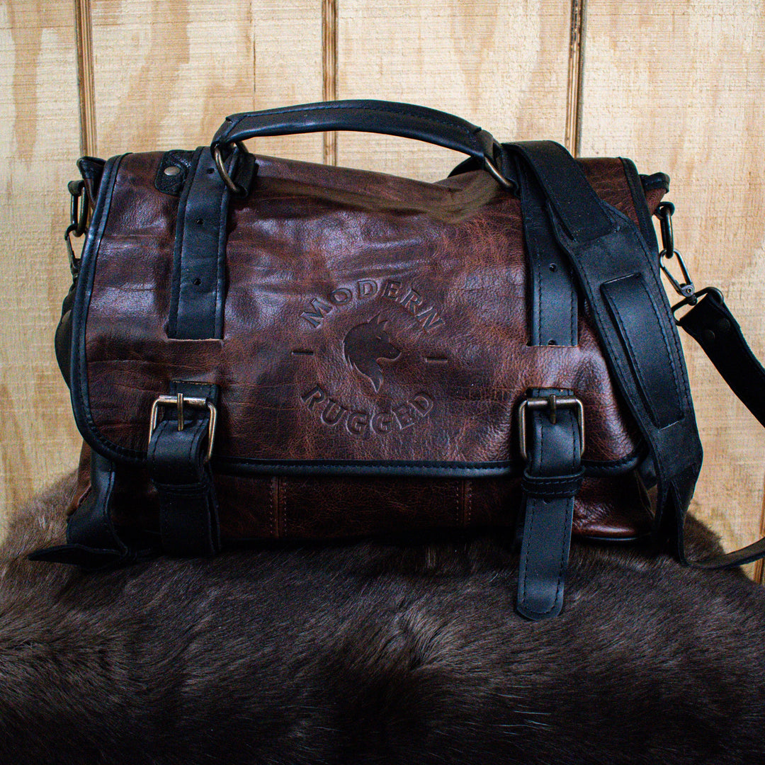 A leather hide Modern Rugged laptop bag with brass zippers and buckles, primarily in rustic merlot and black colors, displayed on a wooden surface.