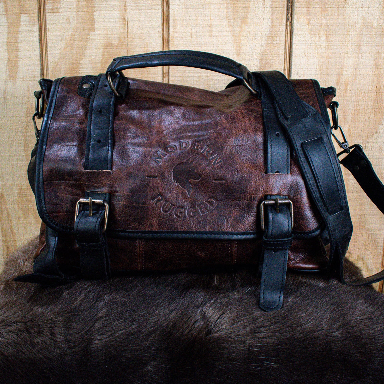 A leather hide Modern Rugged laptop bag with brass zippers and buckles, primarily in rustic merlot and black colors, displayed on a wooden surface.