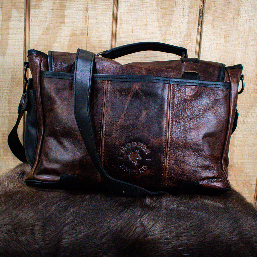 A leather hide Modern Rugged laptop bag with brass zippers and buckles, primarily in rustic merlot and black colors, displayed on a wooden surface.