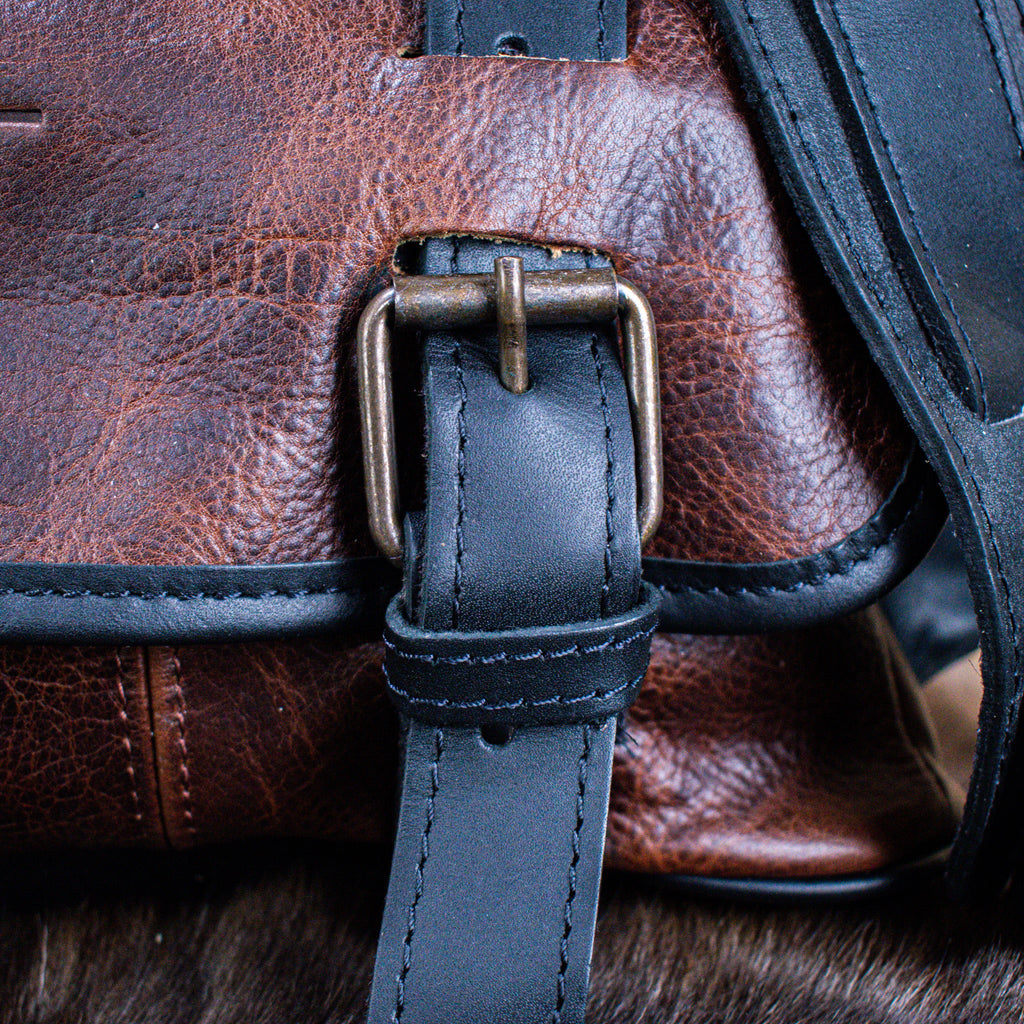 A leather hide Modern Rugged laptop bag with brass zippers and buckles, primarily in rustic merlot and black colors, displayed on a wooden surface.