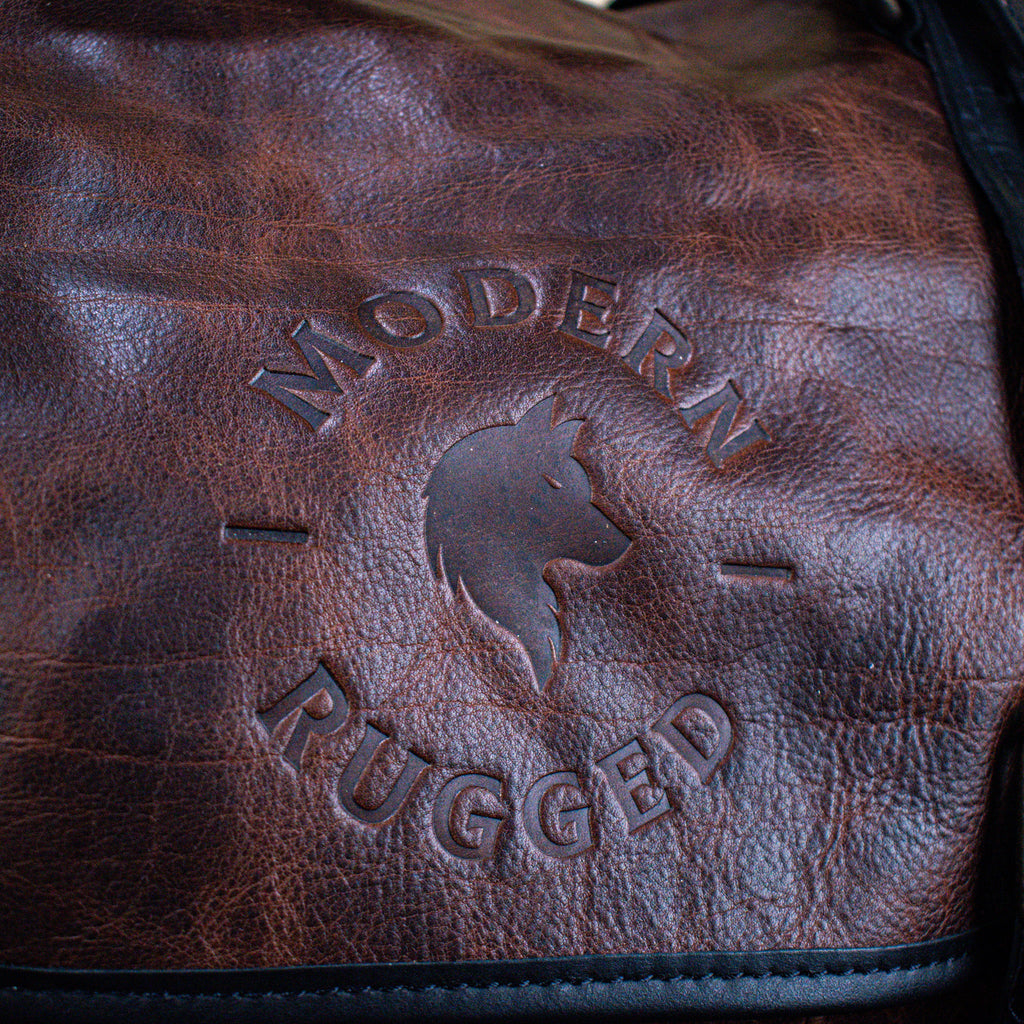 A leather hide Modern Rugged laptop bag with brass zippers and buckles, primarily in rustic merlot and black colors, displayed on a wooden surface.