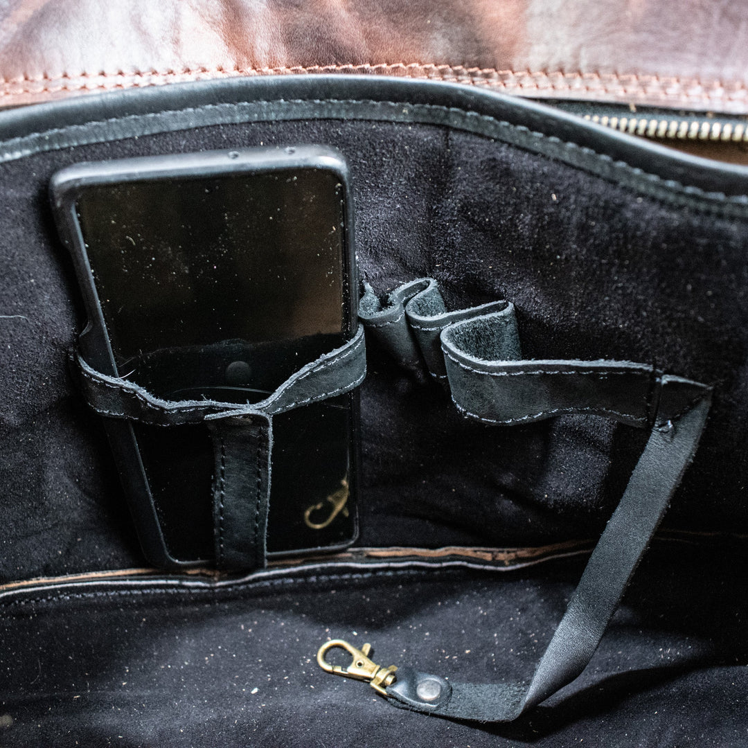 A leather hide Modern Rugged laptop bag with brass zippers and buckles, primarily in rustic merlot and black colors, displayed on a wooden surface.