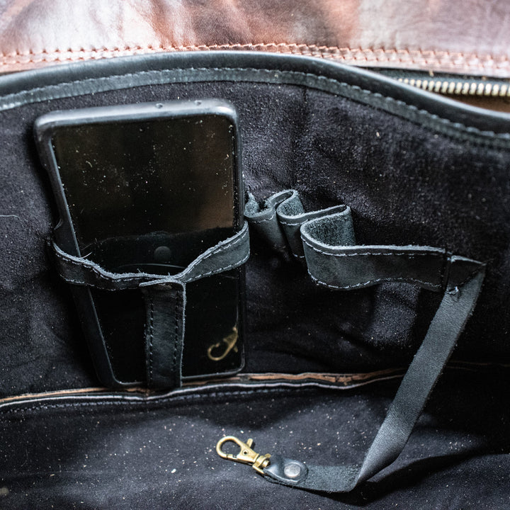 A leather hide Modern Rugged laptop bag with brass zippers and buckles, primarily in rustic merlot and black colors, displayed on a wooden surface.
