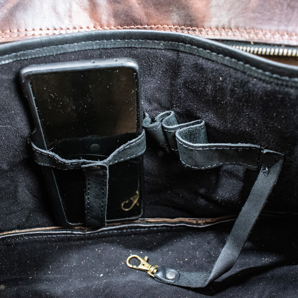 A leather hide Modern Rugged laptop bag with brass zippers and buckles, primarily in rustic merlot and black colors, displayed on a wooden surface.