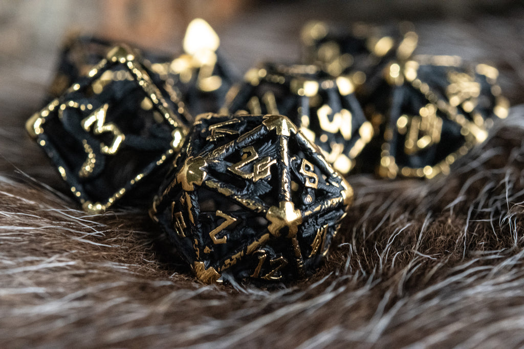 A set of matte black and gold hollow metal dice with embossed numbers, including various shapes such as a d4, d6, d8, d10, d12, and d20, displayed on a white surface.