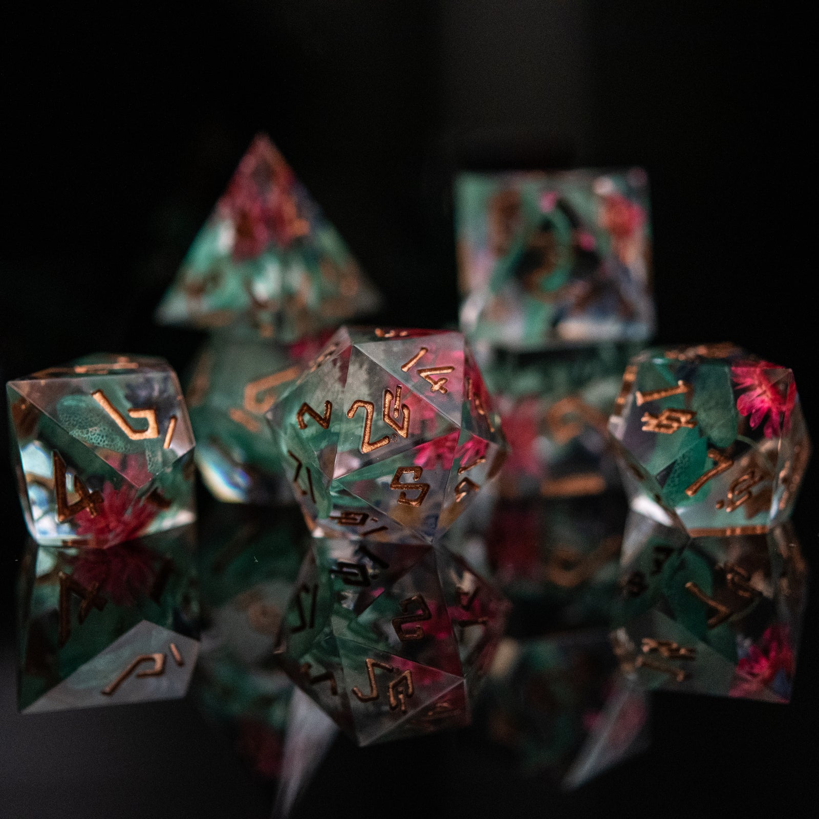A set of sharp-edged resin dnd dice with bronze hand-painted numbers, featuring green and pink botanicals, reflecting on a shiny surface.