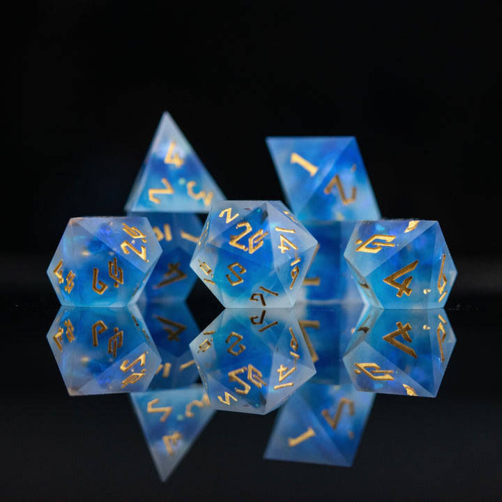 A set of matte blue sharp-edged resin dnd dice with gold engraved numbers, reflecting on a black surface.
