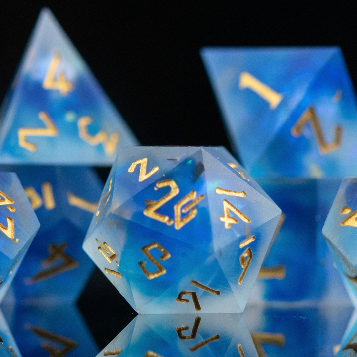 A set of matte blue sharp-edged resin dnd dice with gold engraved numbers, reflecting on a black surface.