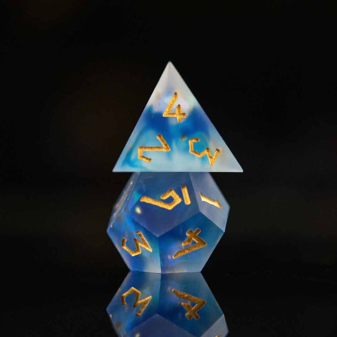 A set of matte blue sharp-edged resin dnd dice with gold engraved numbers, reflecting on a black surface.