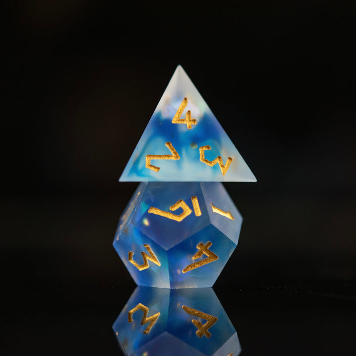 A set of matte blue sharp-edged resin dnd dice with gold engraved numbers, reflecting on a black surface.