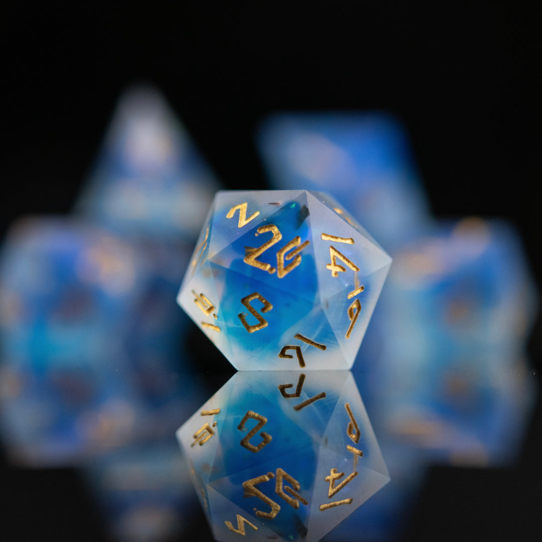 A set of matte blue sharp-edged resin dnd dice with gold engraved numbers, reflecting on a black surface.