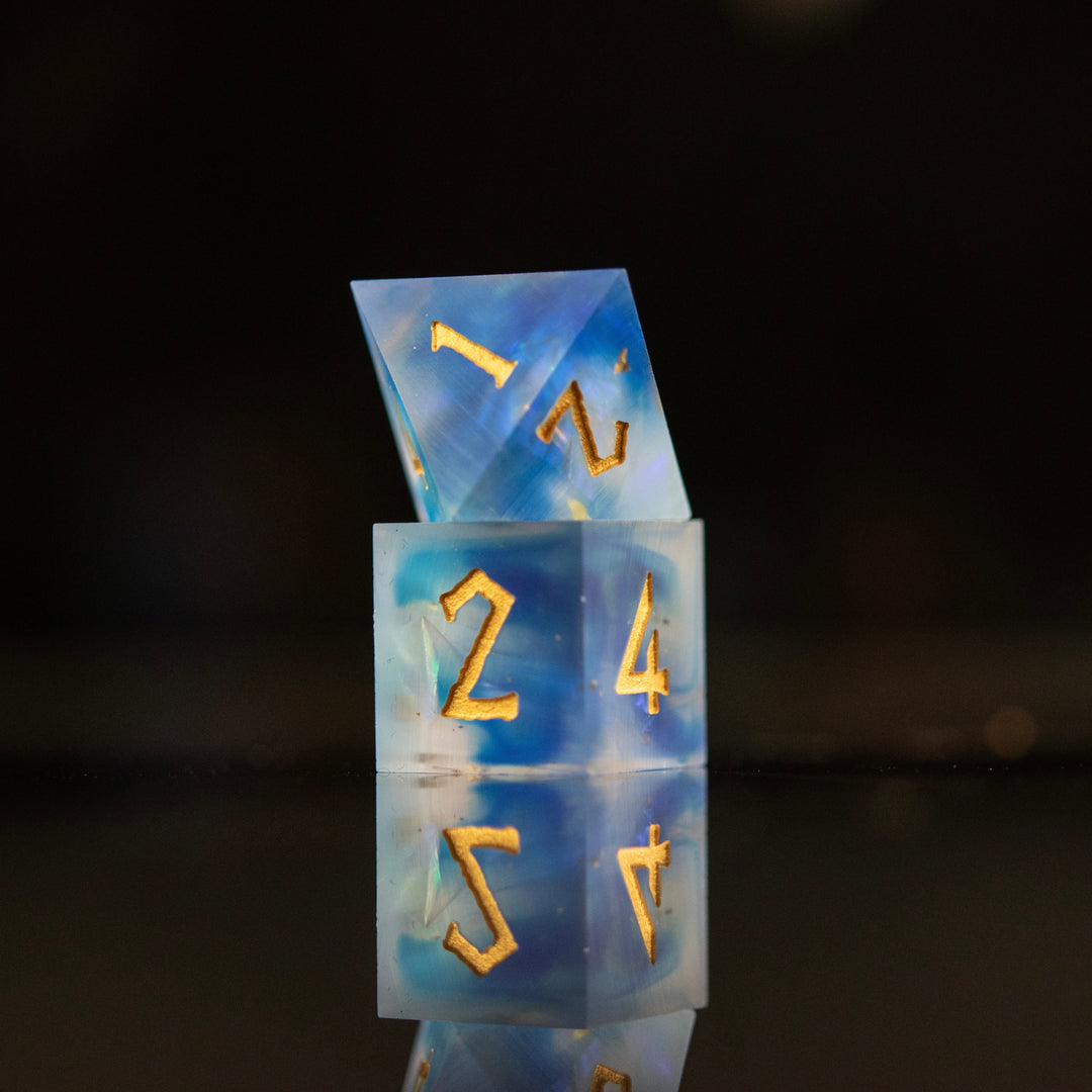 A set of matte blue sharp-edged resin dnd dice with gold engraved numbers, reflecting on a black surface.