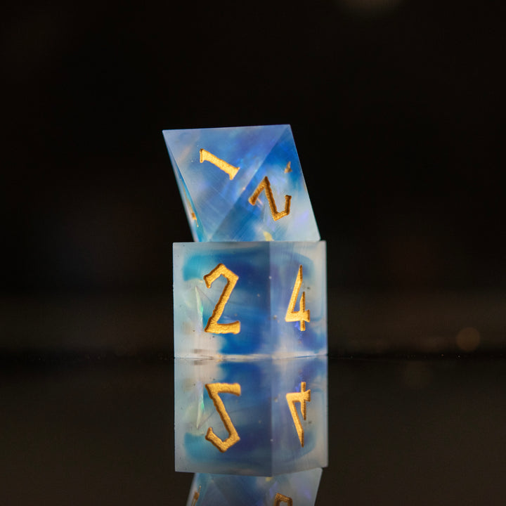 A set of matte blue sharp-edged resin dnd dice with gold engraved numbers, reflecting on a black surface.